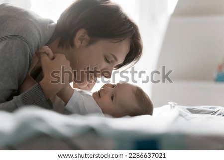 Happy cheerful new mom caressing adorable baby lying on back on bed, kissing sweet child, smiling, laughing, cuddling kid with love, adoration, devotion, affection, enjoying motherhood, parenting