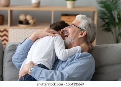 Happy cheerful old senior grandpa embracing cute little boy grandson laughing cuddling at home, two 2 generations family elder grandfather and small grandkid hugging bonding playing sit on sofa