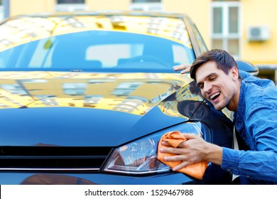 Happy Cheerful Owner Of New Black Car Is Wiping, Cleaning Auto With Orange Rag. Smiling Brunette Man Driver In Denim Shirt Is Hugging And Stroking Vehicle. In Love With Automobile Concept. 