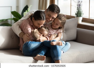 Happy Cheerful Parents Having Fun With Cute Children Daughters Cuddling Playing On Sofa Together, Mom And Dad Laughing Embracing Little Kids Daughters Tickling Enjoying Family Lifestyle Games At Home