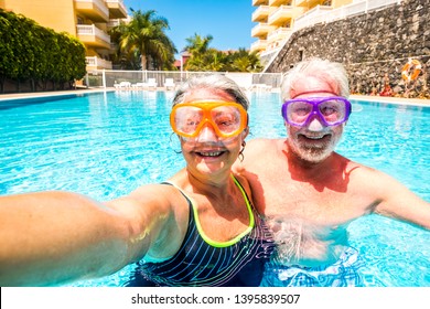 Happy cheerful people old senior man and woman have fun together in the summer swimming pool activity taking selfie pictures with scuba masks on the face for funny outdoor leisure activity in hotel