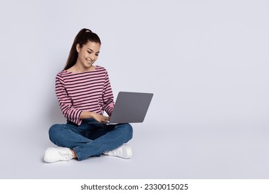 Happy cheerful pretty brunette young woman in casual outfit digital nomad sitting on floor, using modern pc laptop, working online, chatting with friends, shopping, grey background, copy space - Powered by Shutterstock