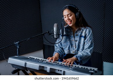 Happy cheerful pretty smiling of young Asian woman vocalist Wearing Headphones recording a song front of microphone and playing the keyboard during rehearsal of her band in a professional studio - Powered by Shutterstock