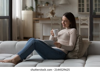 Happy Cheerful Reader Girl Enjoying Novel Story On Couch, Drinking Hot Beverage, Reading Paper Book In Cardboard Cover, Smiling, Laughing, Enjoying Bookworms Hobby, Home Leisure Time