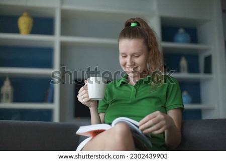 Happy cheerful reader girl enjoying novel story on couch, drinking hot beverage, reading paper book in cardboard cover, smiling, laughing, enjoying bookworms hobby, home leisure time