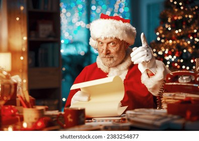 Happy cheerful Santa Claus sitting at his desk, reading letters and pointing with a finger, Chistmas time concept