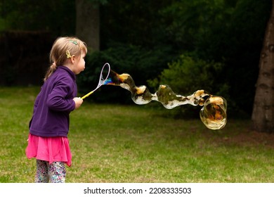 Happy Cheerful School Age Child Blowing Huge Soap Bubbles Outdoors, Young Caucasian Girl Making A Large Soap Bubble Outside In The Park, Alone. Having Fun, Fun Summer Activities People Lifestyle