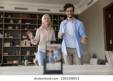 Happy cheerful senior lady and handsome young blogger man dancing at home together, shooting activity, recording video on smartphone, laughing, enjoying online streaming, communication, broadcasting - Powered by Shutterstock