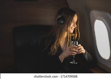 Happy cheerful sexy woman with long blonde hair, in a stylish suit, sitting in an airplane seat, looking out the window, listening to music with headphones, enjoying the flight.