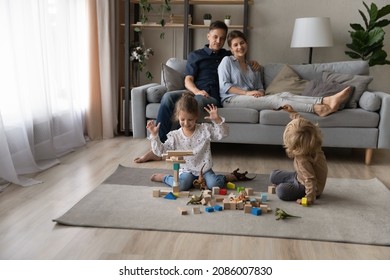 Happy cheerful sibling children playing with building blocks, dinosaurs on warm heating floor, constructing towers. Couple of parents resting on couch, watching sibling kids game with toys