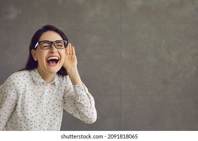 Happy Cheerful Smiling Nosy Beautiful Young Woman In Glasses Puts Hand To Ear Listening To Good Advice, Pleasant Positive News Or Funny Private Information On Gray Marketing Text Copy Space Background