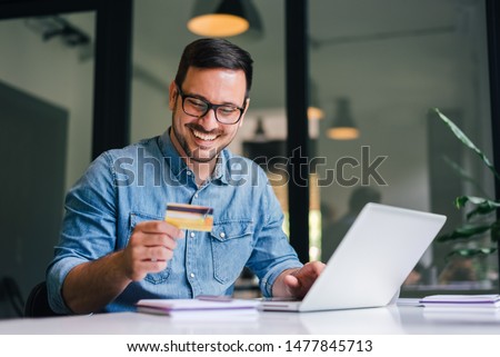 Happy cheerful smiling young adult man doing online shopping or e-shopping satisfied entrepreneur making online payment paying for service or goods self employed freelancer collecting fee