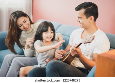 Happy Cheerful Smiling Young Family Mother Father And Daughter Sitting On The Blue Sofa, Father And Daughter Play A Guitar, Rose Pink Color Background Wall 