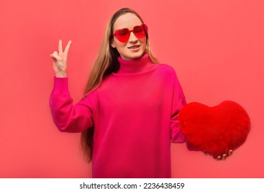 Happy cheerful smiling young woman showing peace gesture v sign with two fingers, wearing pink knitted dress with red trendy heart shaped glasses and holding toy heart on colored pink background.