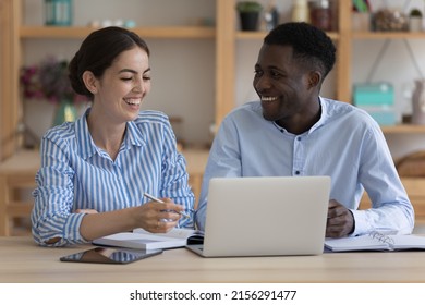 Happy Cheerful Two Diverse Employees Sharing Laptop At Workplace, Talking, Discussing Project, Laughing. Business Team Friends, Interns, Coworkers Watching Learning Video On Computer. Teamwork Concept