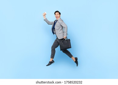 Happy cheerful young Asian businessman jumping while holding bag and coffee cup in isolated studio blue background