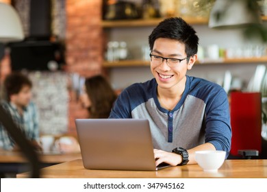 Joyeux jeune homme asiatique dans les lunettes souriant et utilisant son ordinateur portable au café