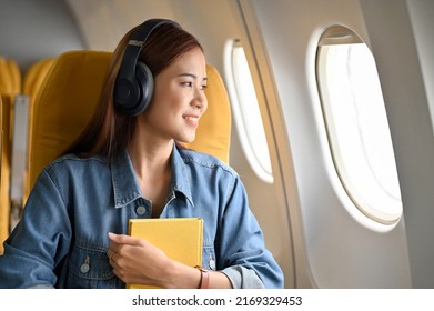 Happy And Cheerful Young Asian Woman With Headphones Looking The View Outside Of The Plane Window. Female Passenger Image