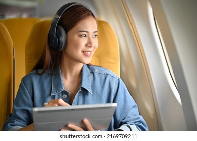 Happy And Cheerful Young Asian Woman With Headphones Using A Portable Tablet And Looking The View Outside Of The Plane Window. Female Passenger Image