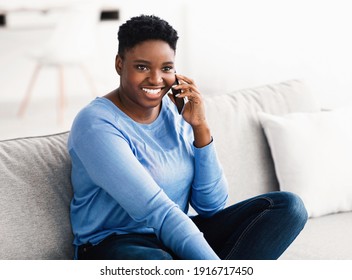 Happy Cheerful Young Black Woman Talking On The Phone At Home, Smiling Lady Making Call By Cellphone Sitting On Sofa At Home, Having Pleasant Or Funny Conversation Speaking By Mobile