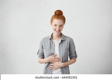 Happy cheerful young Caucasian red haired female with bun smiling cheerfully, keeping hands on her stomach, feeling full after dinner or can't stop laughing at some really good joke. Body language - Shutterstock ID 644581501