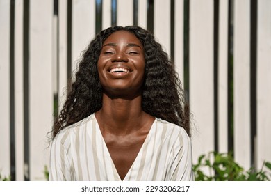 Happy cheerful young ethnic black African attractive pretty woman with curly long hair standing outdoor on sunny city street laughing. Close up portrait, candid authentic shot. - Powered by Shutterstock