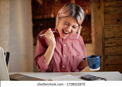 Happy Cheerful Young Female Sitting At Desk With Calculator And Laptop Holding Pen And Mug Having Overjoyed Look, Exclaiming Yes With Excitement, Finally Paid Off Mortgage Debt, Feeling Free