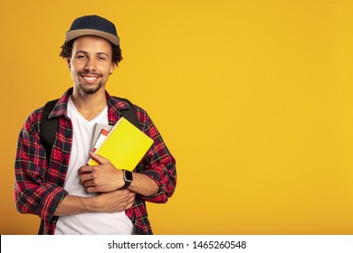 Happy Cheerful Young Latino Student Pose On Camera. Smiling. Hold Yellow Book In Hand. Standing Alone. Isolated Over Yellow Background