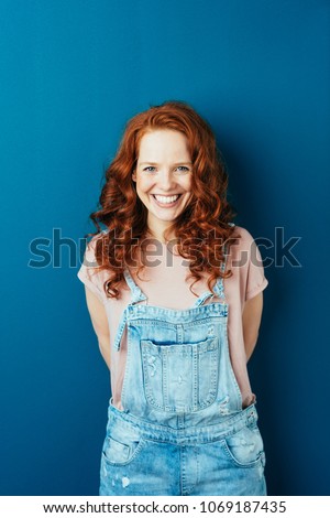 Happy cheerful young redhead woman in dungarees standing over a dark studio background with copy space grinning at the camera