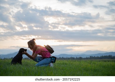 Happy Cheerful Young Woman Kneeling Down To Cuddle Her Cute Black Labrador Puppy As They Are Outside On A Walk In Green Meadow.