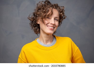 Happy cheerful young woman with short curly hair wearing bright yellow sweater, natural makeup over grey background - Shutterstock ID 2033780885