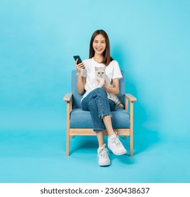 Happy cheerful young woman sitting on the chair and using smartphone with holding a small white cat on blue background. - Powered by Shutterstock