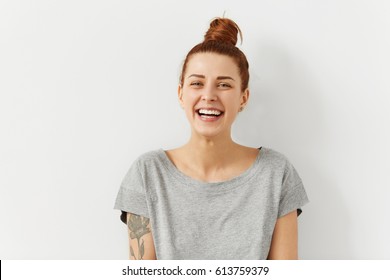 Happy cheerful young woman wearing her red hair in bun rejoicing at positive news or birthday gift, looking at camera with joyful and charming smile. Ginger student girl relaxing indoors after college