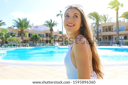 Happy cheerful young woman in white swimsuit and sunglasses on head looking side up next to swimming pool in summer vacation in resort hotel