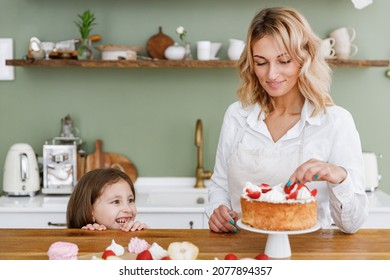 Happy Chef Cook Baker Mom Woman In White Shirt Work Baby Girl Helper Decorate Hide Behind Table At Kitchen Table Home. Cooking Food Process Concept Mommy Little Daughter Prepare Fruit Sweet Pie Cake