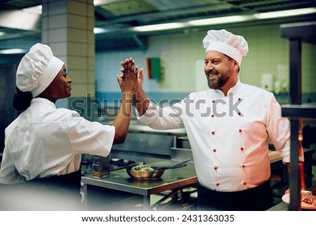 Happy chef giving high five to his African American cook assistant while working in the kitchen.