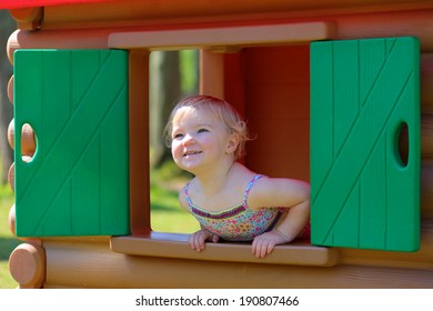 Happy Child, Adorable Blonde Toddler Girl, Having Fun Outdoors Hiding In Plastic Playhouse In Playground On A Sunny Summer Day