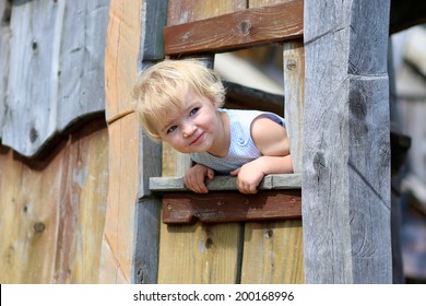 Happy Child, Adorable Blonde Toddler Girl, Having Fun Outdoors At The Park Hiding In Wooden Playhouse In Playground On A Sunny Summer Day