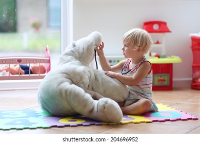 Happy child, adorable blonde toddler girl, playing doctor game with her teddy bear sitting comfortable on the floor in playroom at home, school or kindergarten