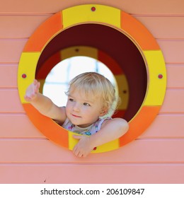 Happy Child, Adorable Blonde Toddler Girl, Having Fun Outdoors Hiding In Plastic Playhouse In Playground On A Sunny Summer Day