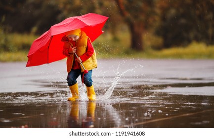 Happy Child Baby Boy With Rubber Boots And Umbrella Jump In Puddle On An Autumn Walk
