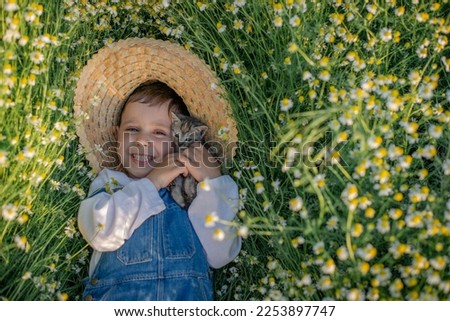 Happy child boy in a straw hat with a cute kitten lies on a lawn of daisies