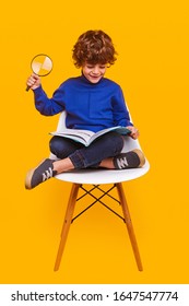 Happy Child In Casual Clothes Smiling And Using Magnifying Glass To Read Book While Sitting Crossed Legged On Chair