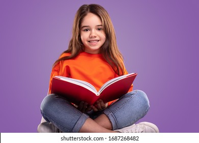 Happy Child In Casual Clothes Smiling And Looking At Camera While Sitting Crossed Legged Against Violet Background And Reading Book During School Lesson