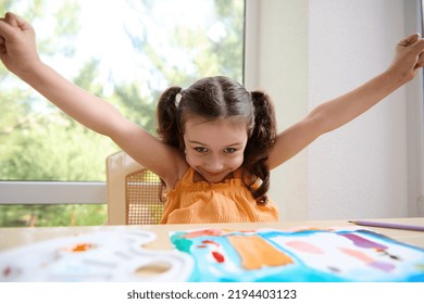 Happy Child, Caucasian Little Preschool Age Girl With Two Funny Ponytails, Raising Her Arms While Sitting At A Table With Drawn Pictures And Cutely Smiles Looking At The Camera, Enjoying Art Class