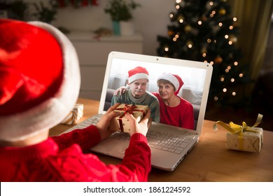 Happy Child  In A Christmas Hat With A Christmas Gift Using A Laptop Communicates With Their Grandparents On Video Call Using Webcam.  Stays At Home For Christmas Holidays.