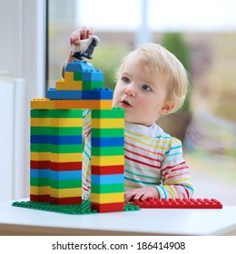 Happy Child, Cute Blonde Toddler Girl Building House From Plastic Blocks Sitting Next To A Big Window Indoors At Home Or Kindergarten
