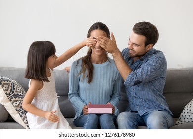 Happy child daughter and husband congratulating mom celebrating birthday at home together closing eyes of smiling mum holding pink gift box, kid and dad make surprise present on mothers day holiday - Shutterstock ID 1276178980