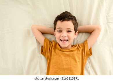 Happy Child With Funny Face. Portrait Of Caucasian Brunette Boy. 7-year-old Kid In A Yellow T-shirt Put His Hands Behind His Head, Lying On Bed. Cotton Pastel Linen Of Olive Color On The Background