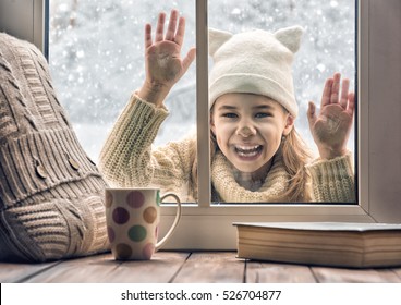 Happy Child Girl Playing On A Winter Walk In Nature. Cute Girl Looking In Window, Standing Outdoors On Winter Forest Background. 